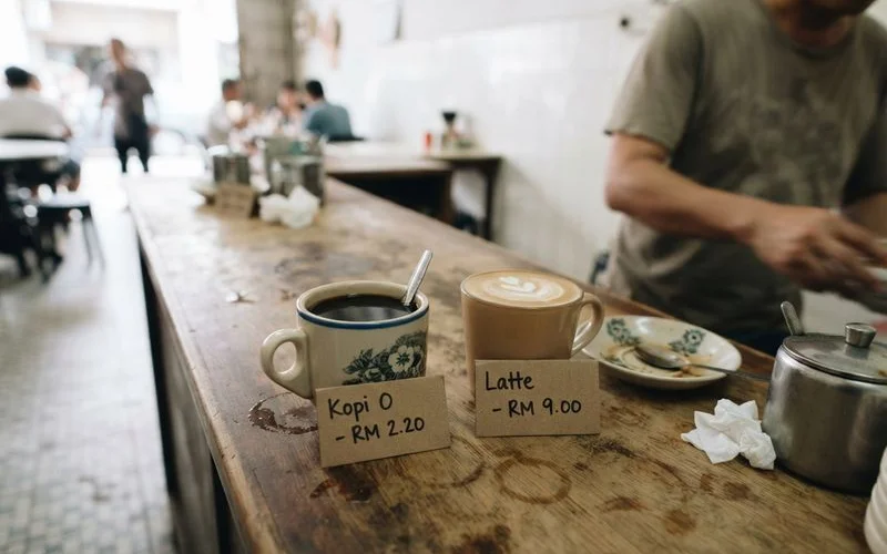 Side by side comparison of a traditional kopitiam coffee cup and a specialty pour-over coffee setup on a wooden counter